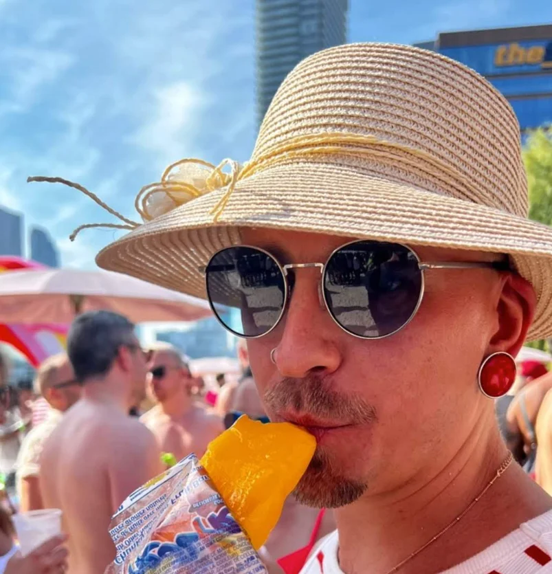 Me, Benjie Kibblewhite, a man, wearing a fetching sun hat, nice red earrings, sunglasses, eating a popsicle with a cheeky tilt to my head.